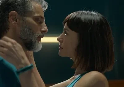 Homem de barba grisalha e mulher de cabelo curto se olham intensamente. Mãos dela no pescoço dele, em cenário escuro e íntimo.