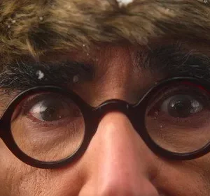 Close-up de rosto com óculos redondos e chapéu de pele nevado. Olhar intenso. Clima frio e misterioso.