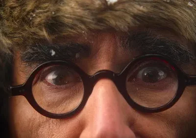 Close-up de rosto com óculos redondos e chapéu de pele nevado. Olhar intenso. Clima frio e misterioso.