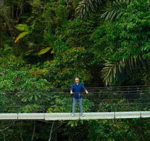 Homem de camisa azul em ponte suspensa alta sobre selva tropical densa. Ele olha para frente, em cenário de exploração.