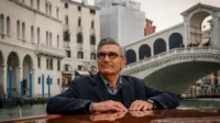 Homem de óculos, cabelo grisalho, em um barco em Veneza, olhando para o lado. Ao fundo, um canal tranquilo, edifícios e a Ponte de Rialto.