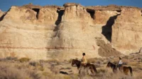 Dois cavaleiros atravessam um deserto com falésias imponentes. Aventura e exploração em paisagem árida sob céu azul.