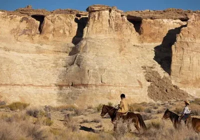 Dois cavaleiros atravessam um deserto com falésias imponentes. Aventura e exploração em paisagem árida sob céu azul.