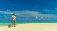 Homem com camisa floral e mala em praia paradisíaca, olhando um hidroavião no céu azul. Cenário de viagem ou isolamento.
