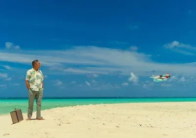 Homem com camisa floral e mala em praia paradisíaca, olhando um hidroavião no céu azul. Cenário de viagem ou isolamento.