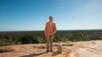Homem de casaco bege e óculos, com maleta, em topo rochoso. Vastidão verde sob céu azul, clima de aventura solitária.
