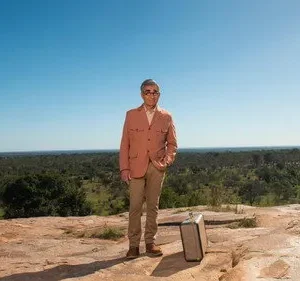 Homem de casaco bege e óculos, com maleta, em topo rochoso. Vastidão verde sob céu azul, clima de aventura solitária.