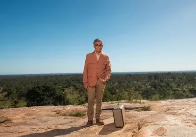 Homem de casaco bege e óculos, com maleta, em topo rochoso. Vastidão verde sob céu azul, clima de aventura solitária.