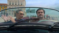 Dois homens em conversível. O mais velho gesticula, o jovem dirige sério. Fundo urbano com prédios históricos e cais ensolarado.