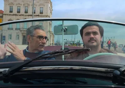 Dois homens em conversível. O mais velho gesticula, o jovem dirige sério. Fundo urbano com prédios históricos e cais ensolarado.