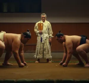 Dois lutadores de sumô ajoelhados no ringue. Um juiz com roupa tradicional os observa. Tensão num cenário sombrio.