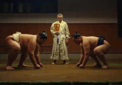 Dois lutadores de sumô ajoelhados no ringue. Um juiz com roupa tradicional os observa. Tensão num cenário sombrio.