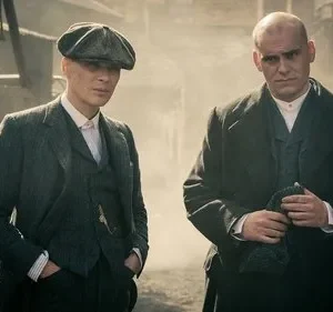 Dois homens sérios, um de boina e terno, outro careca de casaco, em cenário industrial sombrio. Atmosfera tensa.