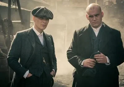 Dois homens sérios, um de boina e terno, outro careca de casaco, em cenário industrial sombrio. Atmosfera tensa.