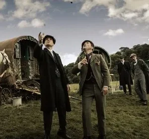 Homens de época em campo, dois olham para o céu, um aponta. Caravana, cavalo branco e carro antigo. Cenário rural e vintage.