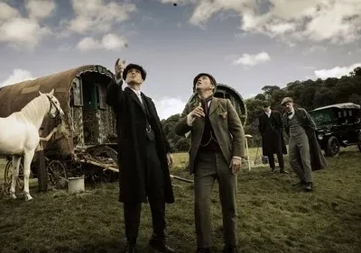 Homens de época em campo, dois olham para o céu, um aponta. Caravana, cavalo branco e carro antigo. Cenário rural e vintage.
