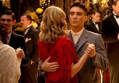 Casal elegante dança em festa dos anos 20/30. Ele de terno risca de giz, ela de vestido vermelho. Outros convidados e músico ao fundo.