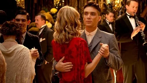 Casal elegante dança em festa dos anos 20/30. Ele de terno risca de giz,ela de vestido vermelho. Outros convidados e músico ao fundo.