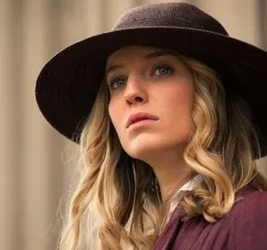 Mulher loira de olhos azuis com chapéu marrom e jaqueta vinho, olhar sério. Fundo bege e desfocado.