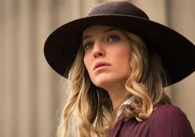 Mulher loira de olhos azuis com chapéu marrom e jaqueta vinho, olhar sério. Fundo bege e desfocado.