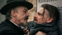 Dois homens se encaram tensos. Um de chapéu e barba intimida o outro, que está encostado numa parede com cartazes.