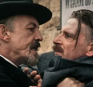 Dois homens se encaram tensos. Um de chapéu e barba intimida o outro, que está encostado numa parede com cartazes.