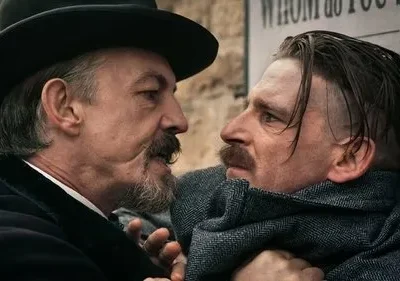 Dois homens se encaram tensos. Um de chapéu e barba intimida o outro, que está encostado numa parede com cartazes.