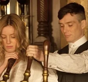 Homem e mulher em roupas de época, em bar vintage. Ambos olham para baixo, sérios, enquanto operam torneiras de chope.