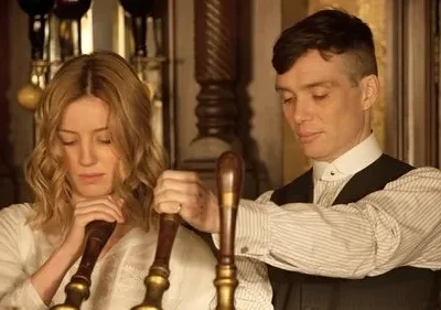 Homem e mulher em roupas de época, em bar vintage. Ambos olham para baixo, sérios, enquanto operam torneiras de chope.