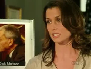 Mulher de cabelo castanho ondulado fala, olhando para a direita, perto de um retrato de um homem grisalho. Cenário profissional e sério.