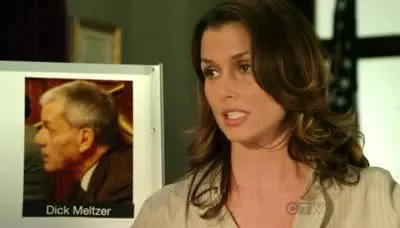Mulher de cabelo castanho ondulado fala, olhando para a direita, perto de um retrato de um homem grisalho. Cenário profissional e sério.