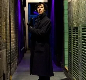 Homem de sobretudo escuro em beco urbano estreito. Paredes de tijolo e azulejos verdes. Luz roxa dá um ar misterioso.