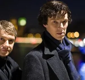 Dois homens em foco. Um sério, de casaco e cachecol azul; o outro sorri discretamente. Noite urbana com luzes desfocadas.