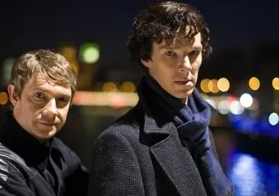 Dois homens em foco. Um sério, de casaco e cachecol azul; o outro sorri discretamente. Noite urbana com luzes desfocadas.