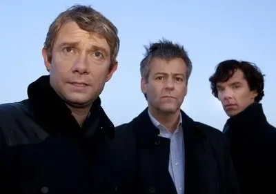 Três homens em casacos escuros. Um preocupado, dois sérios. Posam contra um céu azul pálido. Clima de mistério e investigação.