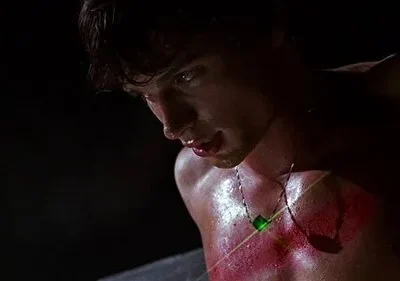 Jovem sem camisa, suado, com marcas vermelhas no peito. Um colar com pingente verde brilhante irradia um laser sobre ele. Olhar intenso.