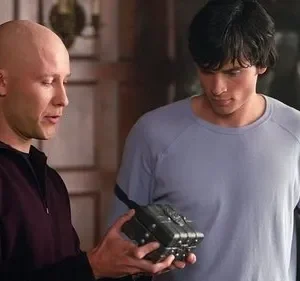 Homem careca e homem de cabelo escuro olham intensamente para uma caixa metálica detalhada. Cenário interno, clima intrigante.