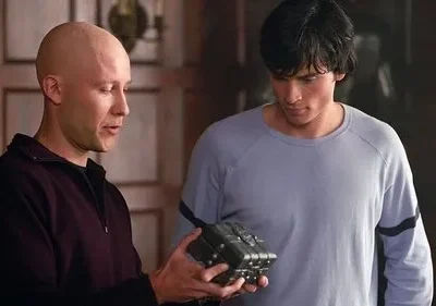 Homem careca e homem de cabelo escuro olham intensamente para uma caixa metálica detalhada. Cenário interno, clima intrigante.