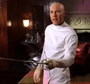 Homem careca com uniforme branco de esgrima e florete, em uma sala luxuosa. Ao fundo, estante com livros e lareira entalhada. Clima formal.