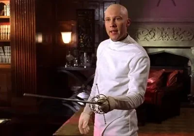 Homem careca com uniforme branco de esgrima e florete, em uma sala luxuosa. Ao fundo, estante com livros e lareira entalhada. Clima formal.