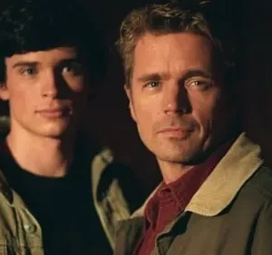 Dois homens sérios. Um jovem de cabelo escuro e um mais velho de cabelo claro, ambos em jaquetas em fundo escuro.