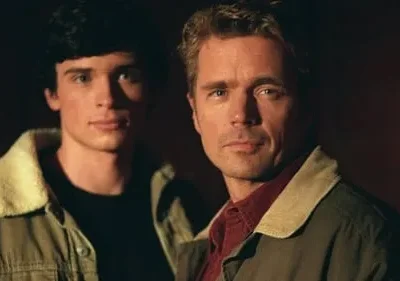 Dois homens sérios. Um jovem de cabelo escuro e um mais velho de cabelo claro, ambos em jaquetas em fundo escuro.