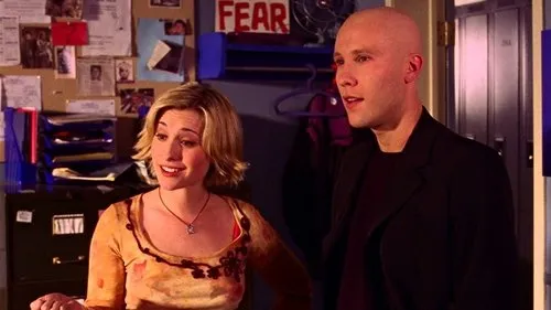 Smallville:As Aventuras do Superboy – T01E07:Desejo 1 Mulher loira sorrindo e homem careca de terno escuro,em um escritório com quadro de avisos e um cartaz FEAR.