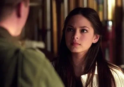 Jovem com cabelo escuro e expressão séria encara um homem de costas. Cenário interno escuro com objetos embaçados.