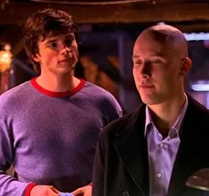 Um jovem de cabelo castanho e camisa roxa olha para um homem careca de casaco escuro. Ambos têm sorrisos sutis em um ambiente escuro.