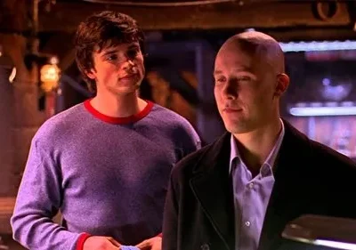 Um jovem de cabelo castanho e camisa roxa olha para um homem careca de casaco escuro. Ambos têm sorrisos sutis em um ambiente escuro.