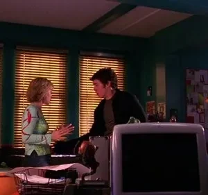 Uma jovem gesticula para um homem num escritório com paredes verde-azuladas e mesas bagunçadas. Luz quente entra por persianas.