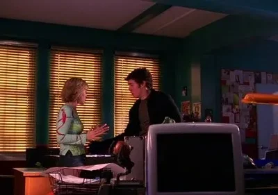 Uma jovem gesticula para um homem num escritório com paredes verde-azuladas e mesas bagunçadas. Luz quente entra por persianas.