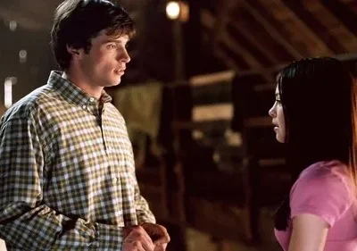 Um homem de camisa xadrez e uma mulher de rosa se encaram intensamente em um ambiente interno escuro de madeira. Clima sério.