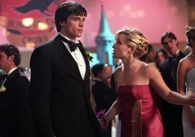 Homem de smoking e mulher de vestido vermelho num baile. Ela o confronta enquanto ele olha distante. Luzes e banner festivo ao fundo.